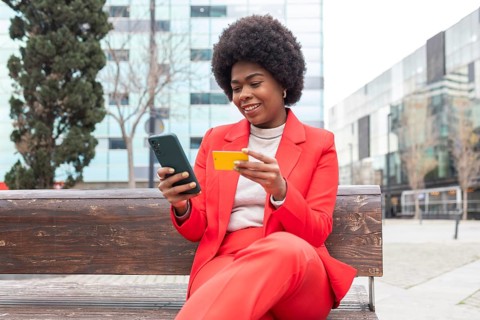 A woman in a vibrant red suit sits on a wooden bench, smiling as she holds a smartphone and a