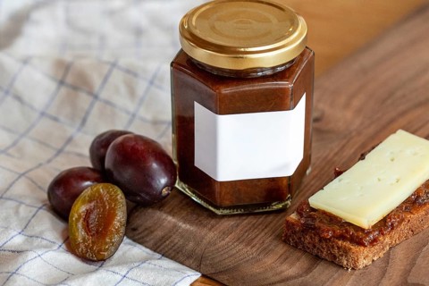 A hexagonal jar of plum compote with a blank label is displayed on a wooden surface. Fresh plums