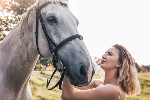 A young woman lovingly interacts with a white horse in a sunlit natural setting. The bond between