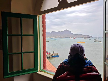 A young woman wearing a braid, cap and backpack in front of a window showing a cloudy sea view and