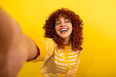 A vibrant, curly-haired woman in a yellow striped sweater joyfully captures a selfie against a