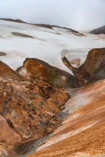 Steaming stream between colourful rhyolite mountains with snowfields, Hveradalir geothermal area,