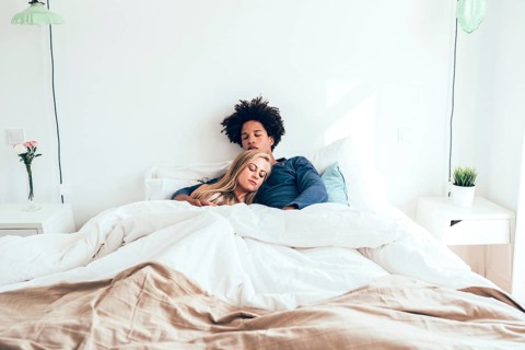 A couple sleeps peacefully on a bed in a minimalist bedroom with white linens and simple decor.