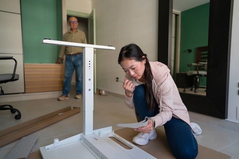 A young Asian woman kneels on the floor, carefully reading the manual while assembling a white desk