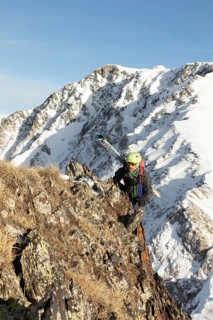 A ski tourer in colorful gear climbs a rugged, snowy mountain ridge under a bright blue sky. The