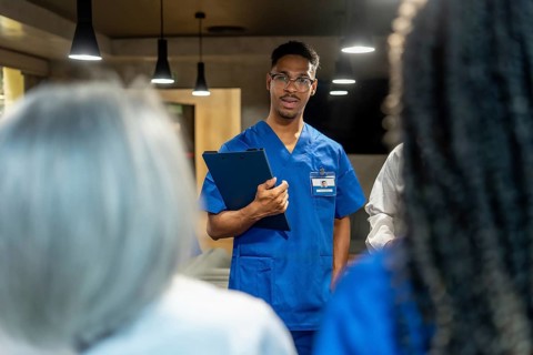 Young black male nurse wearing blue scrubs and glasses holding a clipboard, actively communicating