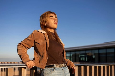 A young mixed-race woman in a brown jacket and jeans stands by a railing, enjoying the sunshine The