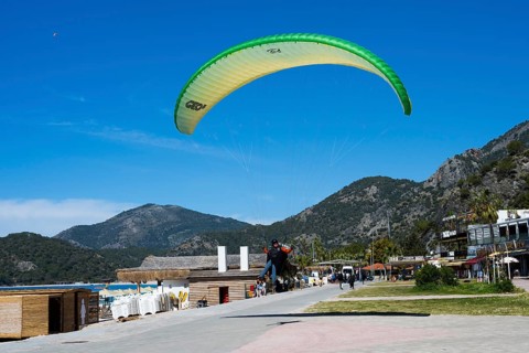 A paraglider floats over a lively beach promenade with mountains in the background, Ölüdeniz,
