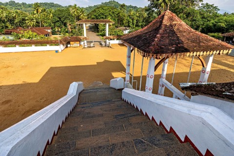 View from a stone staircase of an open sandy square with a traditional pavilion and tropical