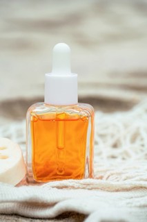 A bottle of hair care oil stands on a sandy textured surface, surrounded by fabric and a loofah.