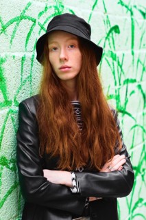 Young woman with long red hair in a stylish black hat and leather jacket poses confidently against