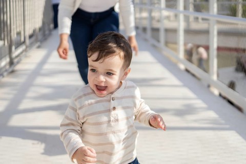 A cheerful baby happily explores the park as his mother follows closely, enjoying a sunny day