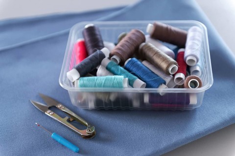 A clear plastic box filled with colorful thread spools sits on folded blue fabric next to small