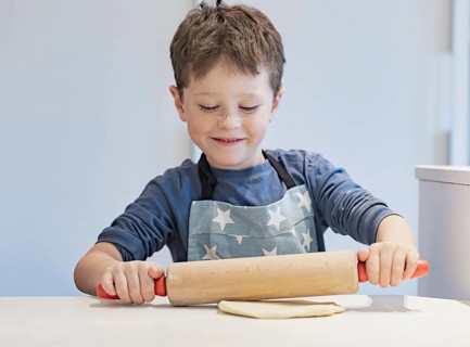 A young child in a star patterned apron happily rolls dough with a wooden rolling pin. His nose is