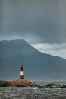 Lonely lighthouse on rocks in the sea with a mountain in the background under a stormy sky, The