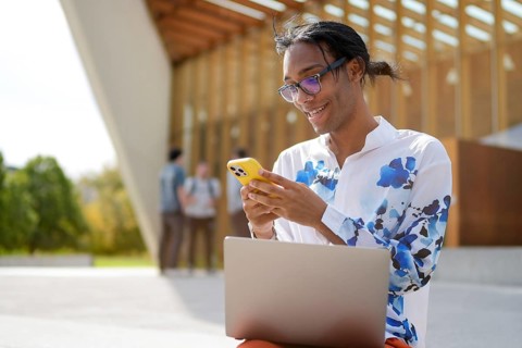 A young man sits outdoors, smiling at his phone while working on a laptop. He is wearing glasses