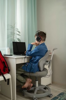 A young boy sits barefoot at a desk, wearing headphones and using a laptop. The desk is in a bright