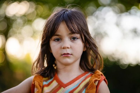 A young girl stands outdoors in a colorful outfit. The background is filled with soft bokeh,