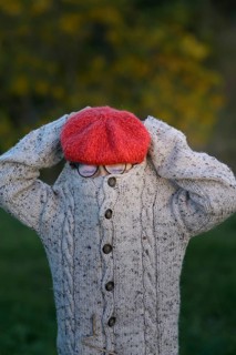 An unrecognizable young boy in a knitted sweater and red beret covers her face, standing in a park