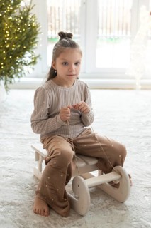 A young girl sits on a wooden toy in a cozy room, decorated with a Christmas tree, during holiday