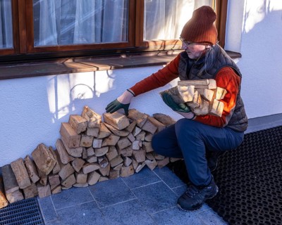 An elderly man in winter attire and gloves carefully arranges eco-friendly firewood outside his