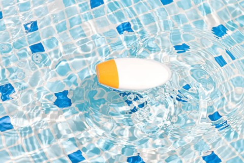 A white and orange cosmetic bottle floats on the clear blue water of a tiled swimming pool,