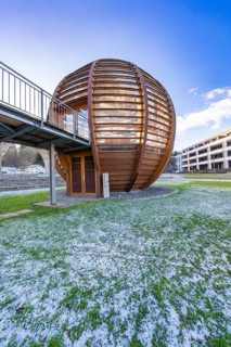 Modern wooden ball with adjoining metal bridge in front of a blue sky, surrounded by buildings and