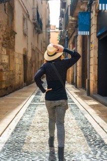 Tourist walking in the medieval city of tarragona, spain, holding her straw hat and looking up at