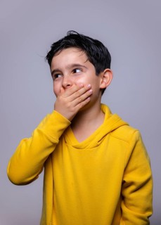 A young boy in a bright yellow hoodie covers his mouth with his hand, glancing sideways with a