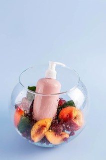 A refreshing pink shower gel bottle sits in a glass bowl surrounded by peaches and berries,