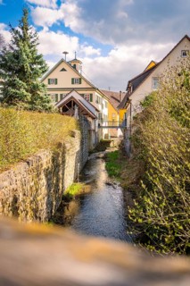 Small river flows between historic houses surrounded by green bushes under cloudy sky, Deufringen,