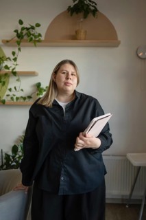 A woman in a black shirt holds a tablet inside a cozy cafe. The setting includes plants and wooden