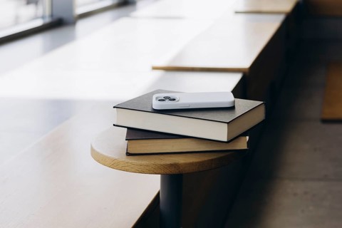 A modern smartphone rests on a stack of two hardcover books placed on a wooden bench. The setting
