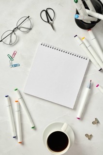 Top view of a sketchbook accompanied by colorful markers, glasses, scissors, paper clips, and a