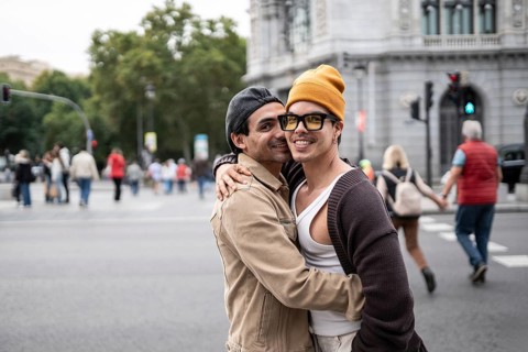 A joyful Colombian gay couple embraces warmly on a bustling city street. They smile brightly,