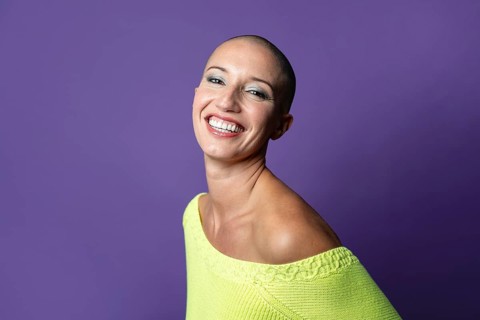 A middle-aged woman with a shaved head smiles confidently against a vibrant purple background,