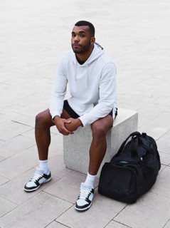 A young black male athlete sitting on a concrete block outdoors. He wears a white hoodie and