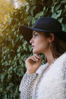 A woman in a black hat, floral-patterned blouse, and white fur vest, gazes thoughtfully against a