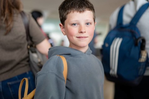 A boy with a backpack stands in a bustling airport terminal. The atmosphere is busy and dynamic,
