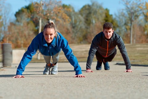 A man and woman exercises outdoors on a sunny day, performing push ups on a concrete path in the