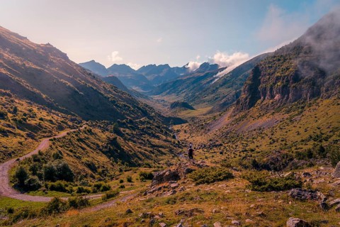 A hiker stands on a rocky outcrop overlooking a majestic mountain valley in the Pyrenees. The scene