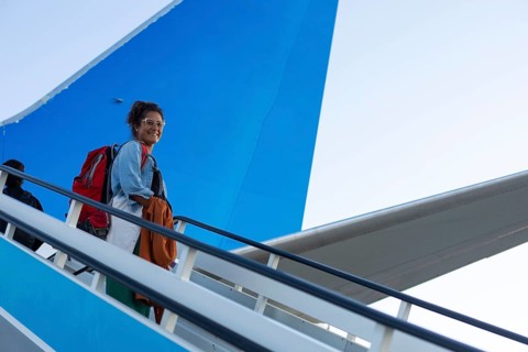 A cheerful traveler ascends airplane stairs with a red backpack against a bright blue sky The scene