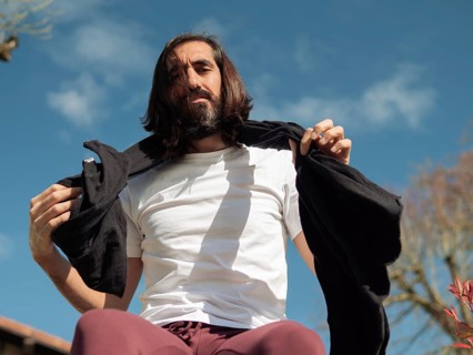 A man with long hair enjoys a sunny day outdoors, wearing a white shirt and holding a jacket. The