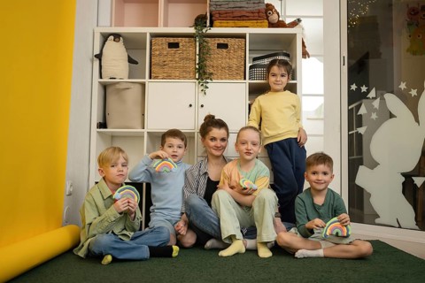 A teacher with five smiling children holding colorful crafts in a classroom. Shelves with