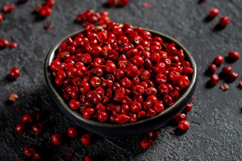 Top view of a dark bowl filled with bright red peppercorns scattered on a textured dark background,