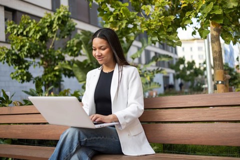 A Hispanic businesswoman in a white blazer works on her laptop while sitting on a bench in a green