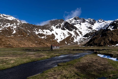 Lonely hut in a wide valley landscape with stream in the foreground and snow-capped mountains in