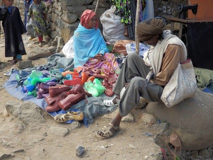 Elderly man wearing traditional clothes tries on shoes at a saleswoman's street stand in Debark,