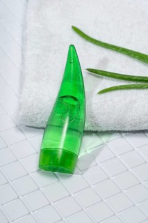 A transparent bottle of aloe vera gel rests on glossy white bathroom tiles alongside a fresh towel