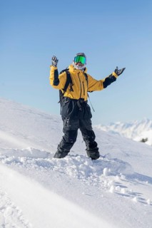 A snowboarder in a yellow jacket enjoys a sunny day on a snowy mountain slope, with clear blue